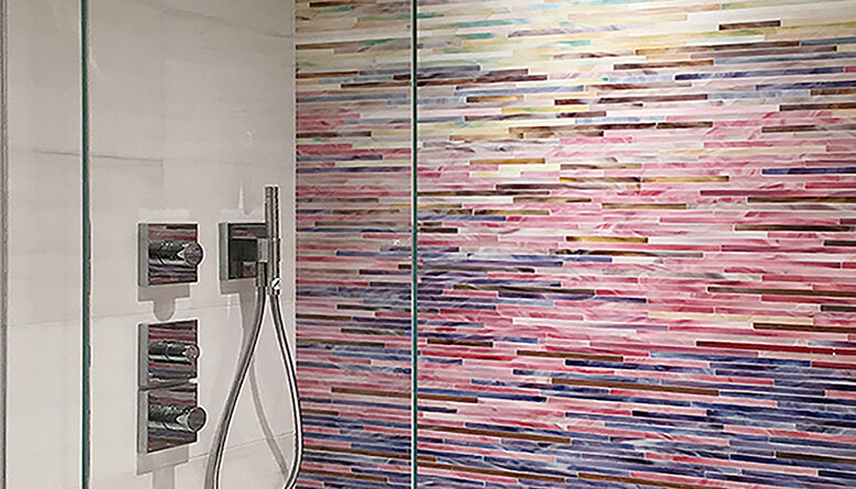 Custom stained glass mosaic shower wall featuring a handcrafted linear gradient in white, pink, blue, bronze, and aqua tones by Allison Eden Studios, paired with white porcelain tile for a luxurious spa-inspired bathroom.