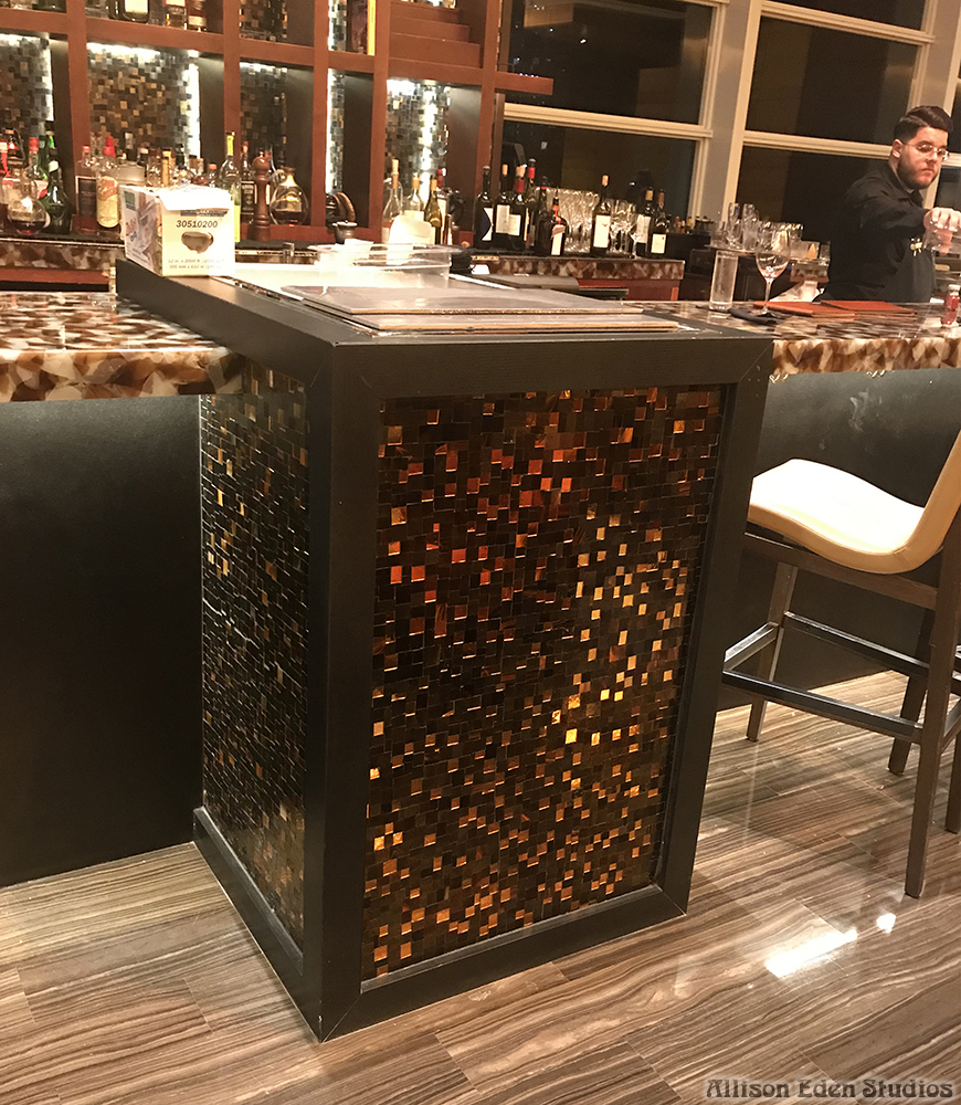 Custom stained glass and mirror mosaic bar backsplash by Allison Eden Studios at the Mandarin Oriental Miami lobby bar featuring illuminated bronze, gold, and metallic glass tiles behind luxury liquor displays close up of stained glass and mirror mosaic hostess stand.