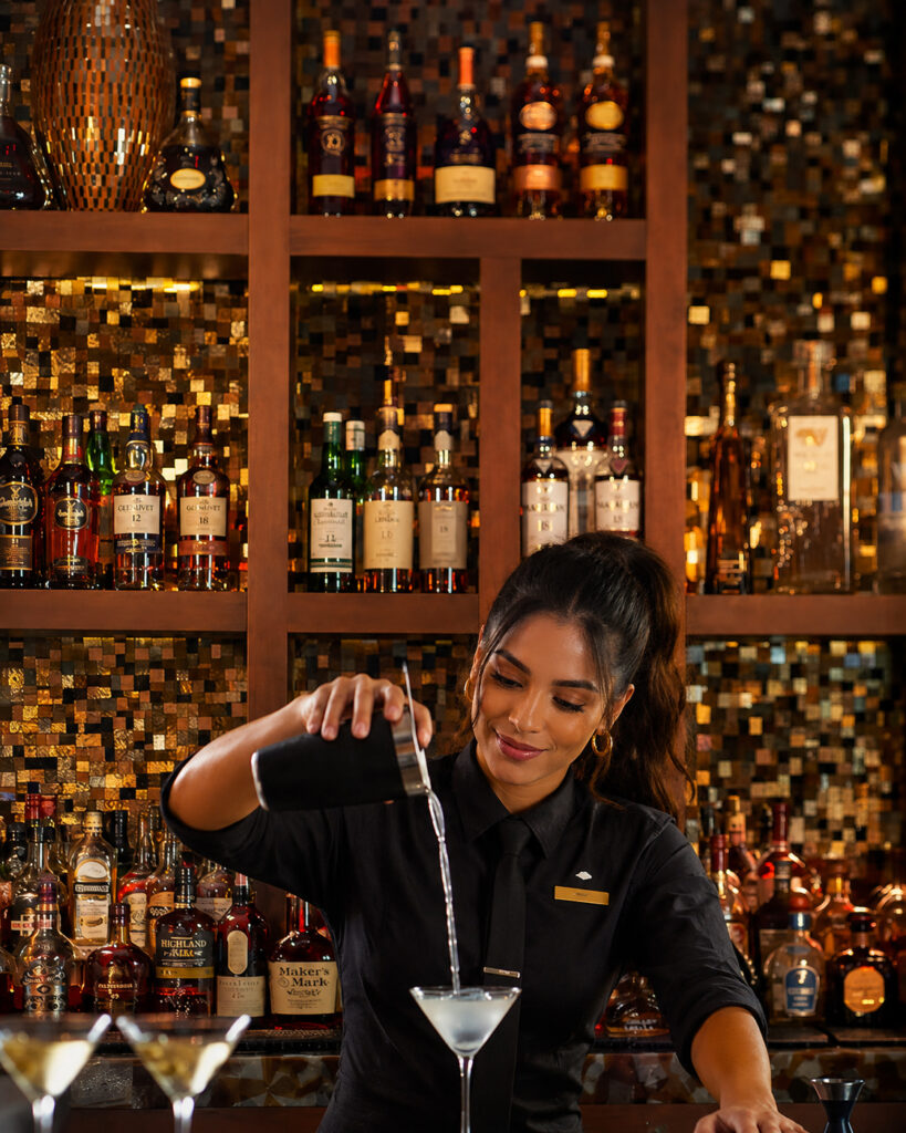 Custom stained glass and mirror mosaic bar backsplash by Allison Eden Studios at the Mandarin Oriental Miami lobby bar featuring illuminated bronze, gold, and metallic glass tiles behind luxury liquor displays with cute bartender.