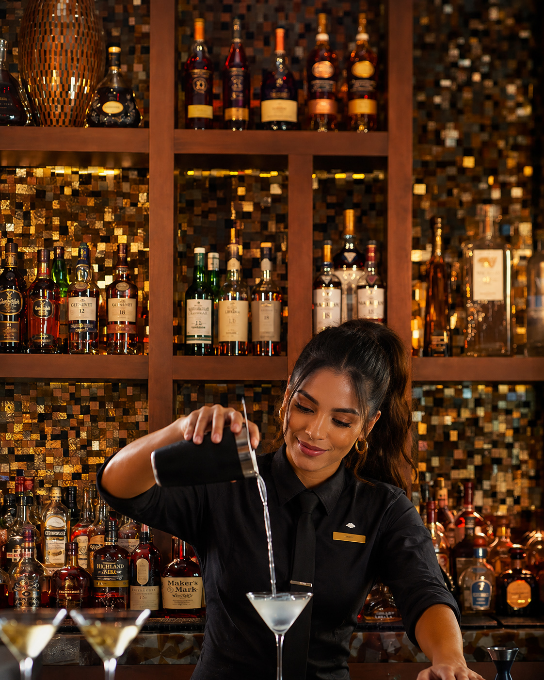 Custom stained glass and mirror mosaic bar backsplash by Allison Eden Studios at the Mandarin Oriental Miami lobby bar featuring illuminated bronze, gold, and metallic glass tiles behind luxury liquor displays cute bartender.