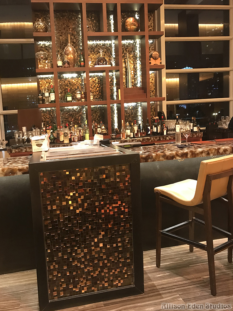 Custom stained glass and mirror mosaic bar backsplash by Allison Eden Studios at the Mandarin Oriental Miami lobby bar featuring illuminated bronze, gold, and metallic glass tiles behind luxury liquor displays with mosaic tile hostell stand.