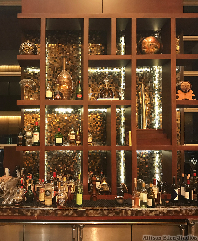 Custom stained glass and mirror mosaic bar backsplash by Allison Eden Studios at the Mandarin Oriental Miami lobby bar featuring illuminated bronze, gold, and metallic glass tiles behind luxury liquor displays shows stained glass and copper mirror backsplash.