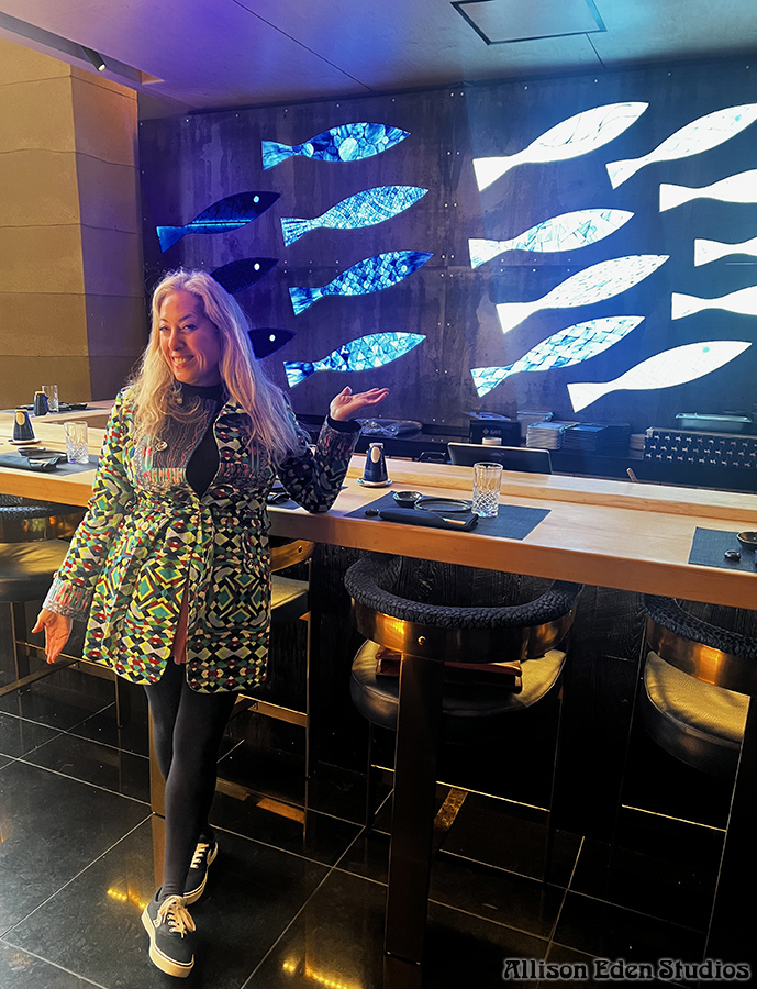 Allison Eden standing beside a custom backlit blue and green stained glass mosaic school of fish at Bondi Sushi in Bryant Park, New York City..