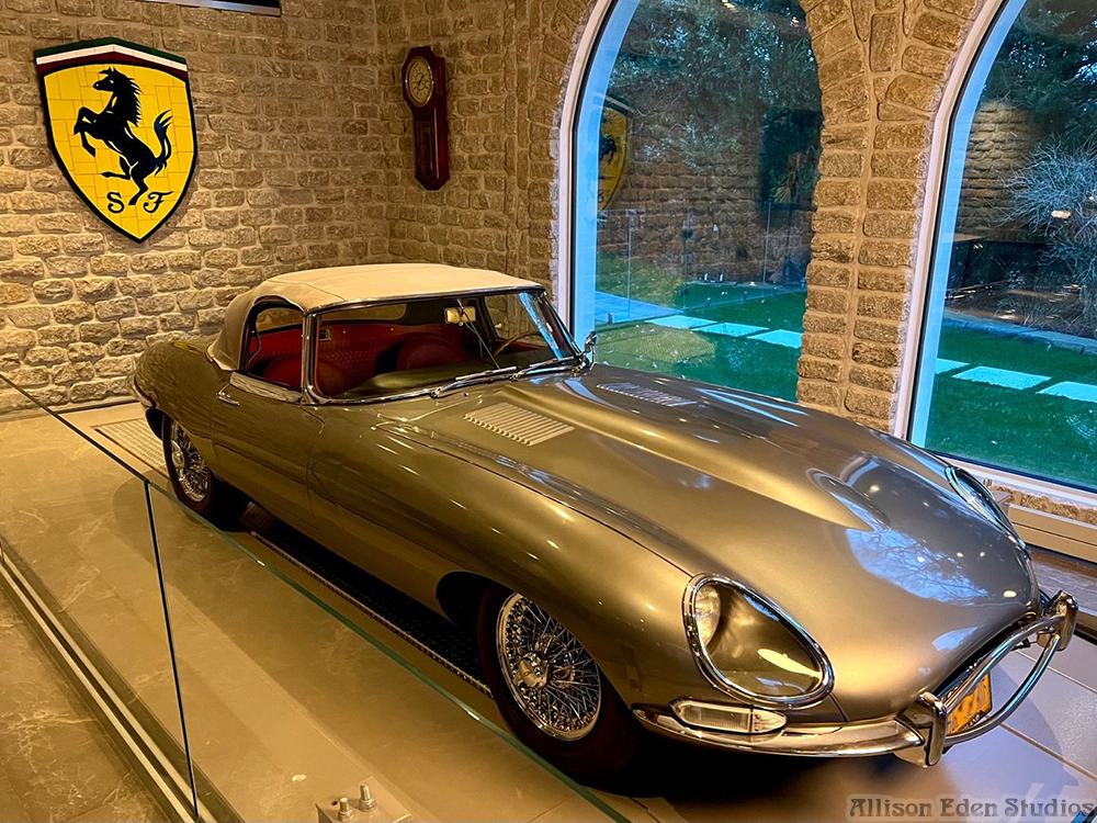 Handcrafted Ferrari-inspired glass mosaic by Allison Eden Studios displayed beside a classic Jaguar E-Type in a private automotive gallery.
