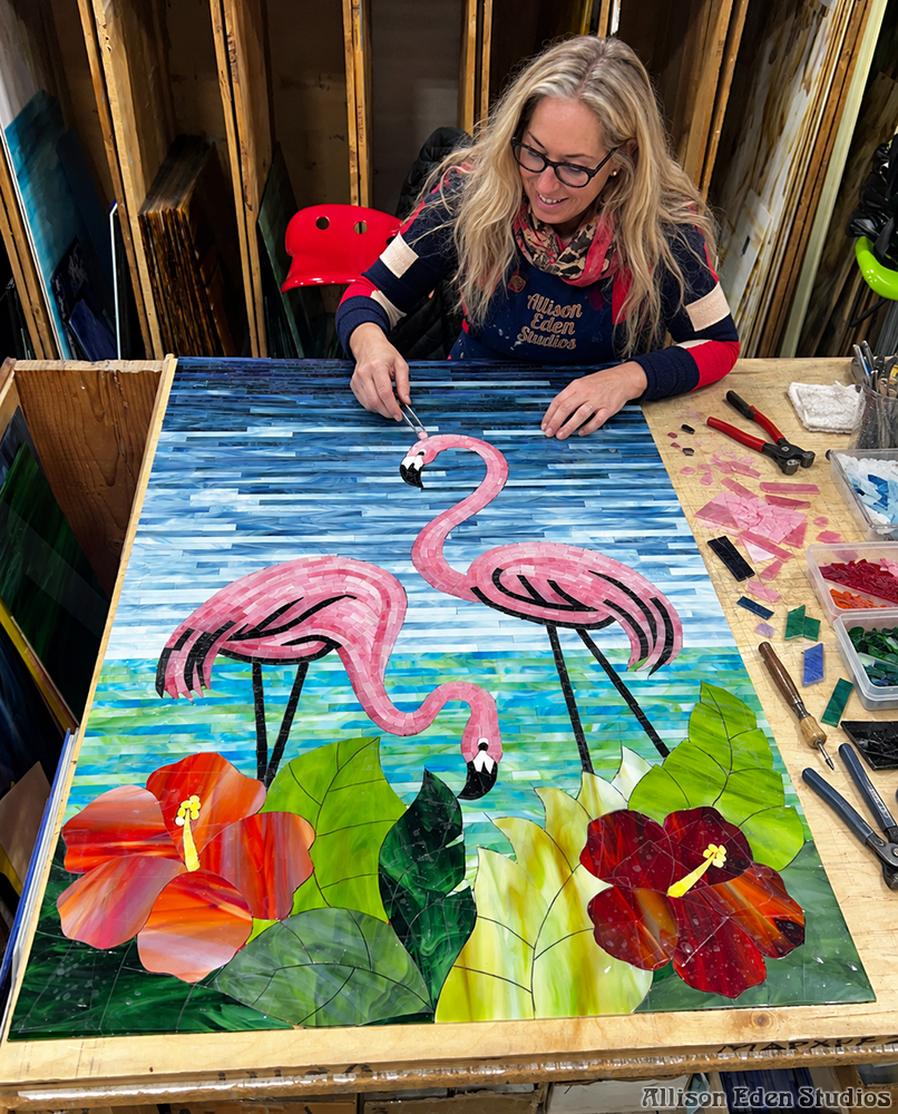 Original tropical stained glass mosaic artwork by Allison Eden featuring two pink flamingos, red hibiscus flowers, green foliage, and a blue ocean background.