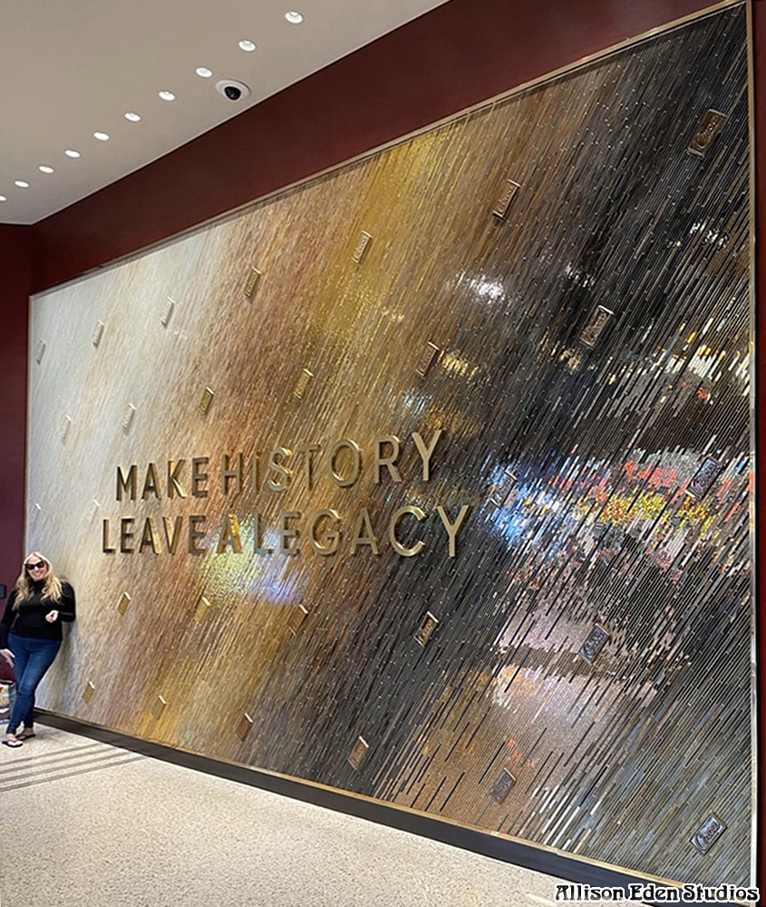 Glass mosaic artist Allison Eden standing beside the monumental Circa Resort & Casino mosaic wall in Las Vegas, showing the scale of the amber and charcoal artwork.