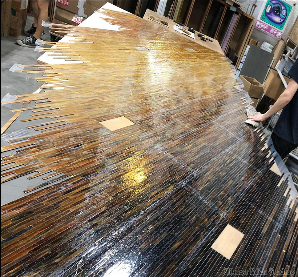 Glass mosaic fabrication in Allison Eden's Brooklyn studio, showing thousands of narrow stained glass pieces arranged in a diagonal bone, amber, chocolate ombre gradation