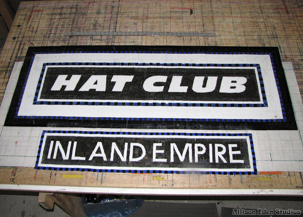 Hat Club and Inland Empire mosaic signs laid out on a workshop table during fabrication, showing precise lettering, cobalt blue borders, and black-and-white glass.