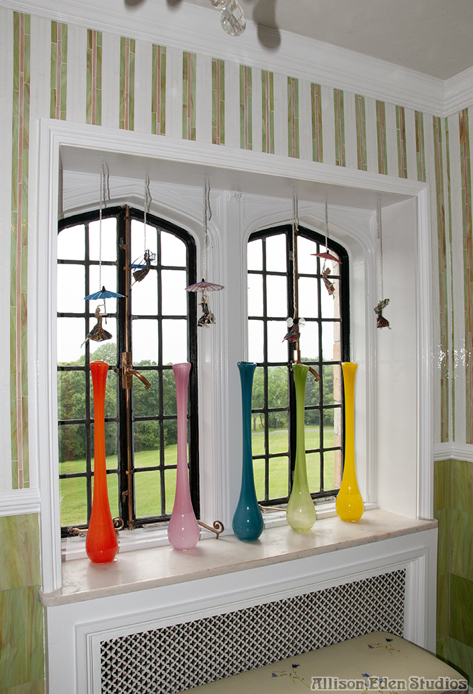 Gatsby-inspired bathroom with pastel green and pink striped glass mosaic walls, white traditional moldings, arched windows, and colorful glass vases in a Mill Neck mansion.