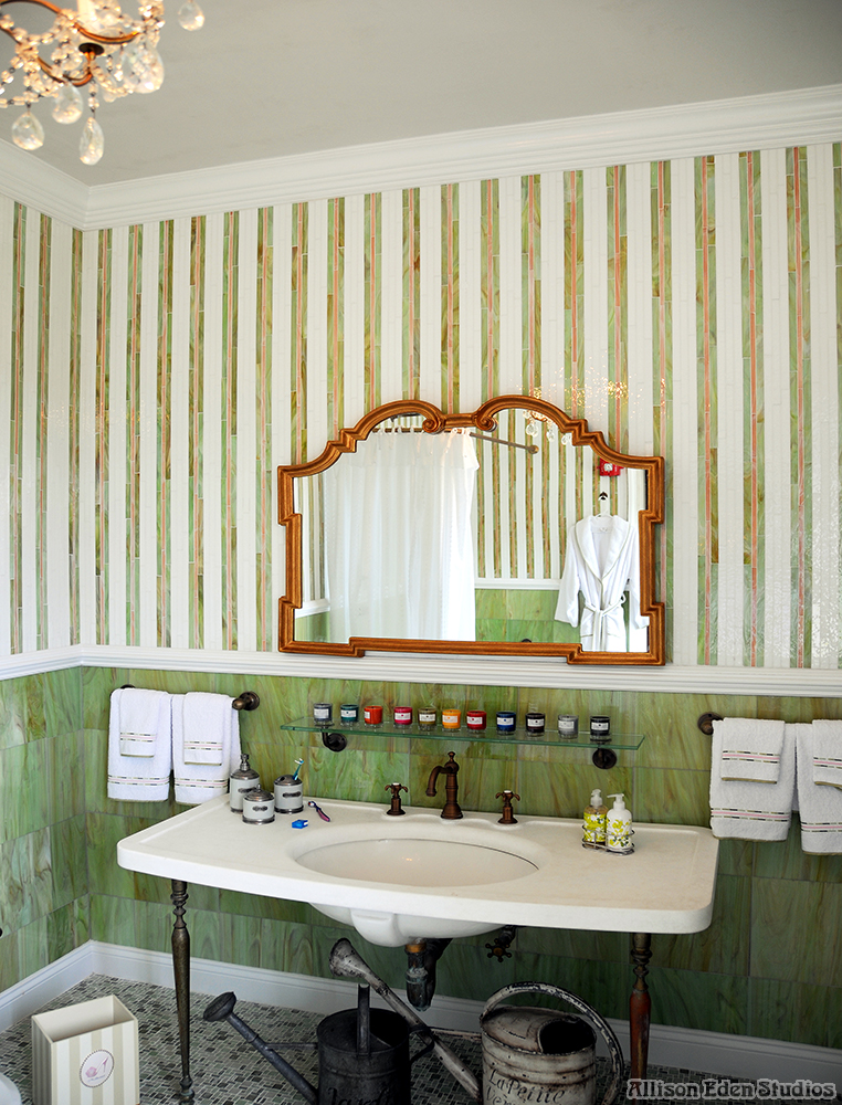 Elegant Gold Coast showhouse bathroom in Mill Neck, NY, featuring Allison Eden's striped glass mosaic walls, green tile wainscoting, ornate gold mirror, and vintage-style sink.