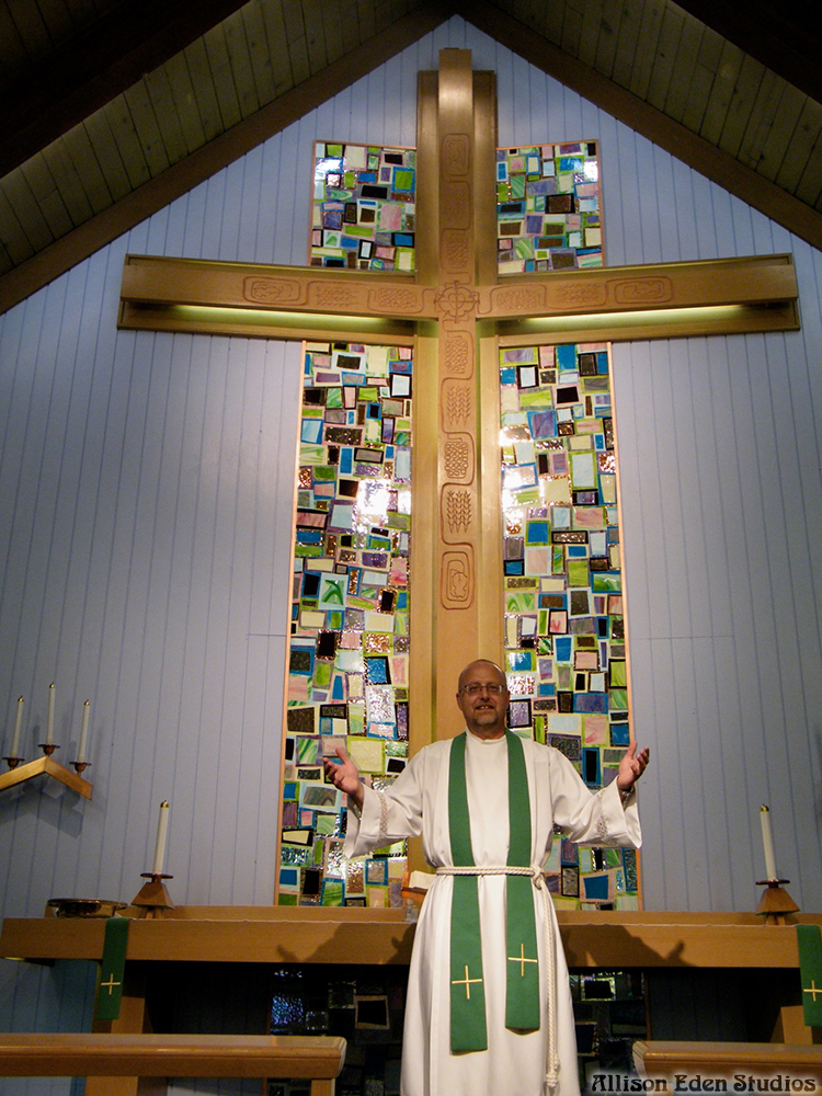 Church pastor standing before a large cross framed by custom stained glass mosaic artwork by Allison Eden Studios.